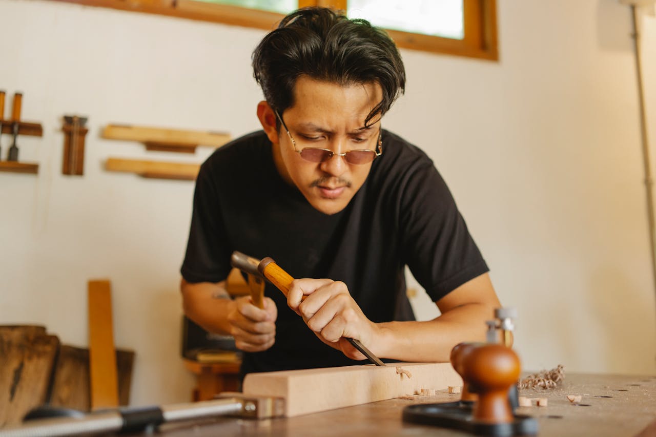 About Concentrated Asian man working with chisel and hammer while cutting notch in hardwood plank
